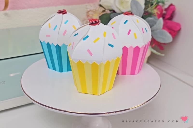 Tre färgglada cupcake-presentaskar i papper på ett vitt tårtfat.